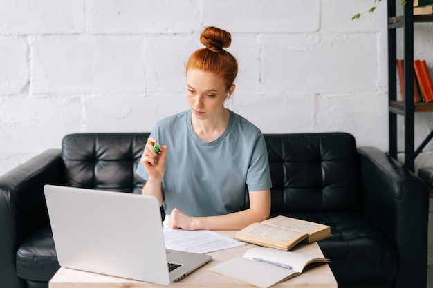 Busy pretty redhead young woman student is taking notes in workbook by looking at laptop screen