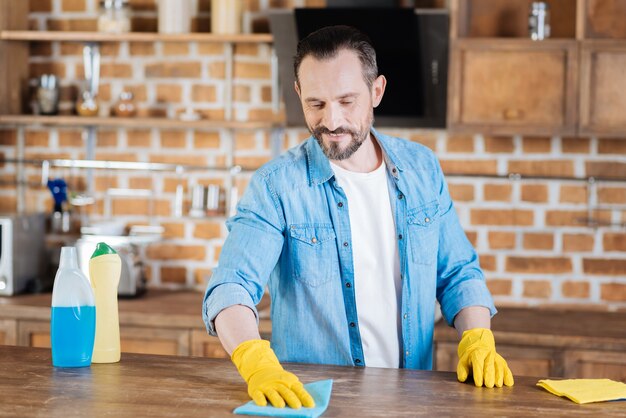 Busy professional male cleaner rubbing from dust while smiling and looking down