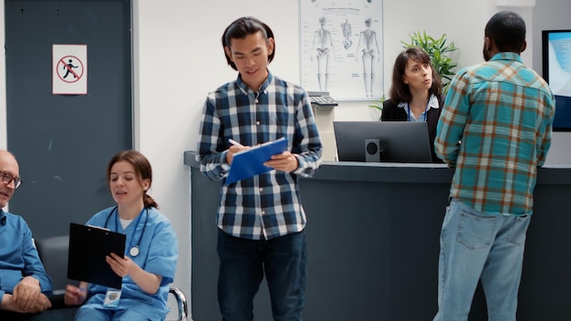 Busy receptionist giving assistance to diverse group of
patients in waiting area lobby, helping with medical reports for
healthcare system. hospital reception desk with many people in
row.