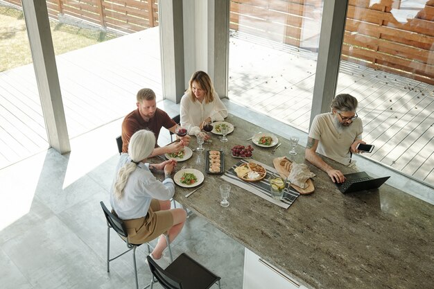 Photo busy senior bearded father in eyeglasses using laptop and talking by phone while working during family dinner