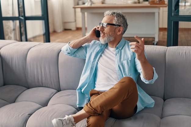 Busy senior man in casual clothing and eyeglasses talking on the smart phone while sitting on the sofa at home