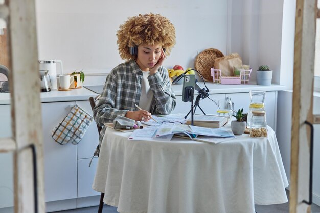 Busy student studies foreign language does exercises on paper\
listens online lecture from tutor wears stereo headphones sits at\
kitchen table prepares for exams blogger records educational\
video