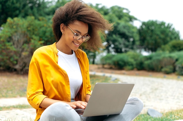 Busy successful african american curly haired young woman student girl freelancer in stylish casual clothes sit with laptop outdoors while online work or study looking for information in internet