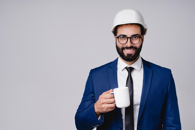 Busy successful young moroccan engineer in hardhat with cup of\
coffee isolated over grey background