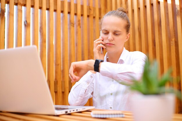 Busy woman wearing white shirt sitting in outdoor cafe working on laptop talking on mobile phone looking at her wristwatch worried about deadline