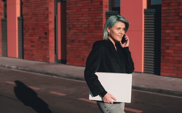 Busy woman with blue hair is walking while holding her laptop and discussing on phone