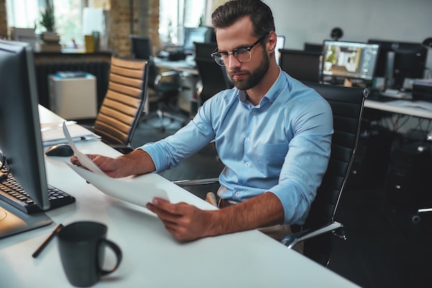 Busy working day concentrated bearded man in eyeglasses and formal wear looking at blueprint while