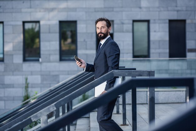 Busy world. Pleasant professional businessman holding his smartphone while walking down the stairs