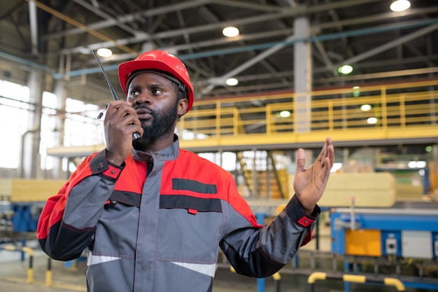 工場でトランシーバーを使用して同僚にタスクを説明しながら手を身振りで示す忙しい若いアフリカ系アメリカ人労働者