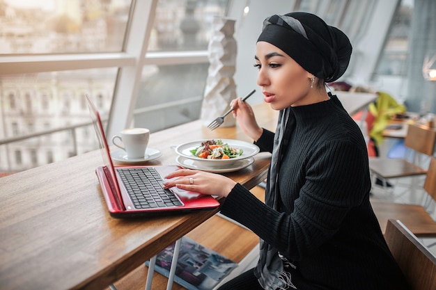 Busy young arabian woman work at laptop and look at it. She sits inside and hold fork upon bowls with food. Model wears hijab.