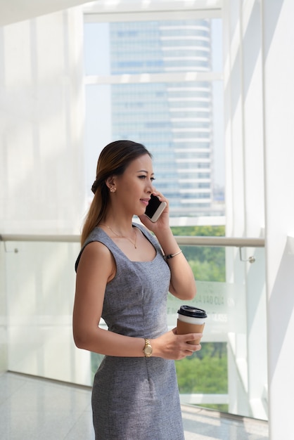 Busy young Asian lady in gray dress standing in office corridor and drinking coffee while answering phone call