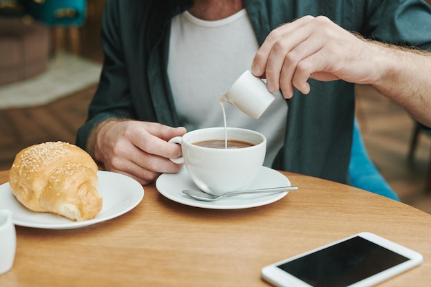 Busy young bearded man sitting at table in modern cafe and drinking coffee while discussing project