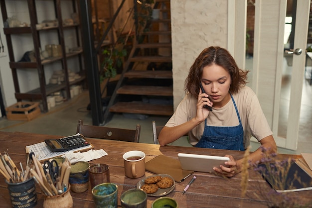 Busy young female potter sitting at desk with tools and ceramic mugs and using tablet while calling ...