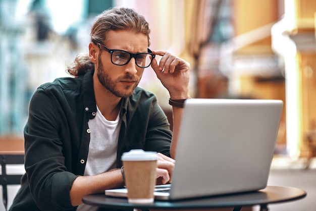 コンピューターを使用して作業し、カフェに座ってコーヒーを飲むカジュアルな服装で忙しい若い男