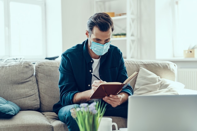 Busy young man in protective face mask writing something down while working from home