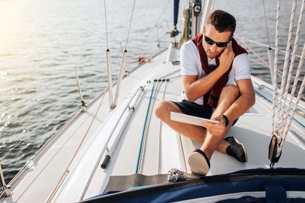 Il giovane occupato si siede a bordo dell'yacht e parla sul telefono. inoltre tiene e guarda tablet. il giovane si siede con le gambe incrociate. indossa occhiali da sole.