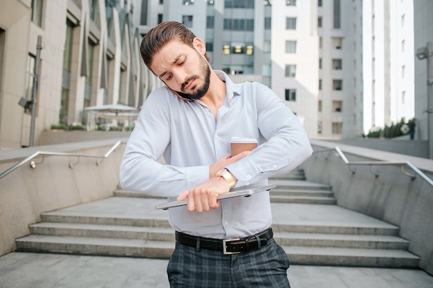 Busy young man stands on steps and looks at watches. He holds laptop, cup of coffee and talks on phone at the same time. Businessman is busy.