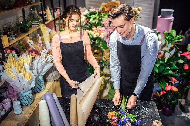 Il giovane e la donna occupati stanno al tavolo. tiene il rotolo di carta e le taglia con le forbici. guy fa il bouquet.