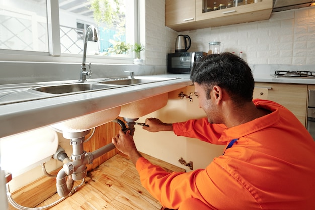 Busy young mixed race plumber in orange bodysuit unscrewing water pipe while changing it under sink in kitchen