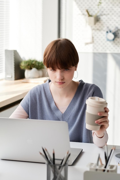 Busy young office woman with short hair using laptop and drinking coffee while working on marketing