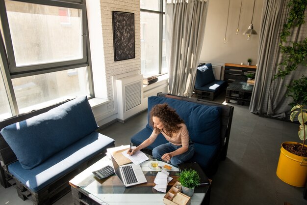 ソファに座ってコピーブックで作業メモを作るカジュアルウェアで忙しい若い女性