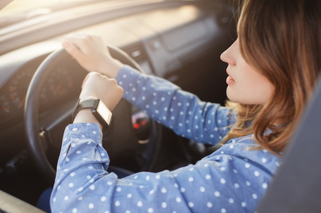 Busy young woman drives car and looks at watch, stuck in traffic jam