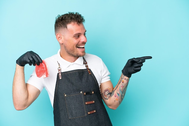 Macellaio uomo caucasico che indossa un grembiule e serve carne fresca tagliata isolata su sfondo blu che punta il dito di lato e presenta un prodotto