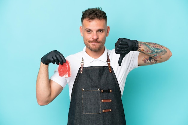 Macellaio uomo caucasico che indossa un grembiule e serve carne fresca tagliata isolata su sfondo blu mostrando il pollice verso il basso con espressione negativa