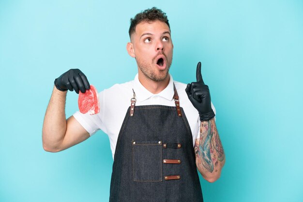 Butcher caucasian man wearing an apron and serving fresh cut meat isolated on blue background thinking an idea pointing the finger up