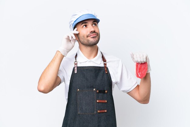 Butcher caucasian man wearing an apron and serving fresh cut meat over isolated white background having doubts and thinking