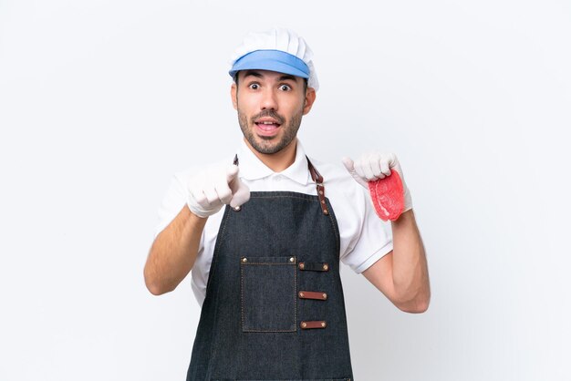 Butcher caucasian man wearing an apron and serving fresh cut meat over isolated white background surprised and pointing front