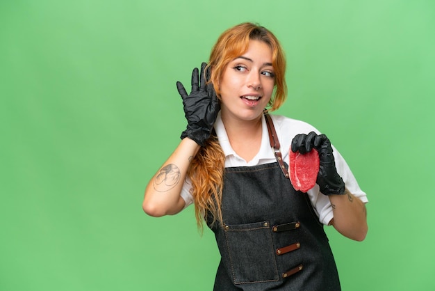 Butcher caucasian woman wearing an apron and serving fresh cut meat isolated on green screen chroma key background listening to something by putting hand on the ear