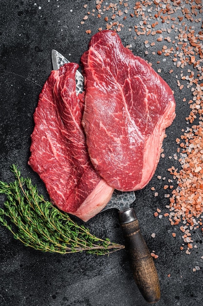 Butcher choise beef raw steaks on butcher knife black\
background top view