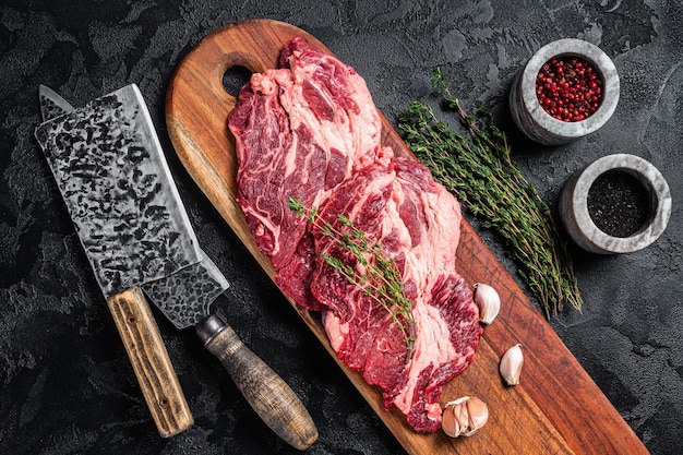 Butcher marbled meat steaks on wooden cutting board raw beef\
ribeye steaks black background top view