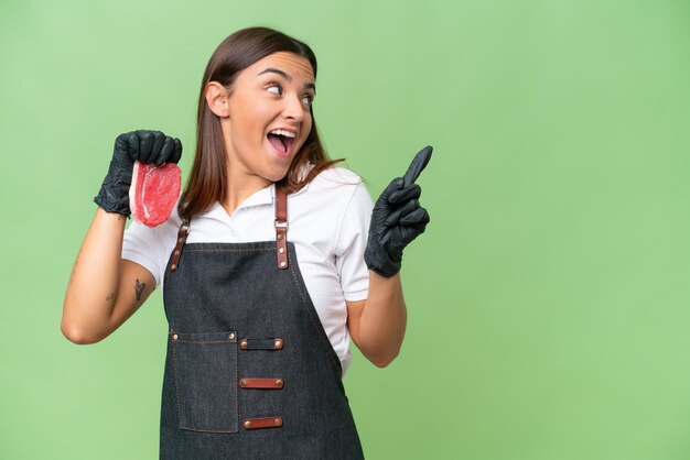 エプロンを着用し、緑の彩度の背景に分離された新鮮なカット肉を提供する肉屋の女性は、指を上げながら解決策を実現しようとしています