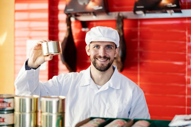 Foto un venditore di macelleria mostra alla telecamera una lattina con carne tagliata e sorride