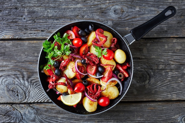 Buttered young potato salad with baby octopuses or moscardini, tomatoes, black olives, and red onion slices, served on a skillet on a wooden table, top view, close-up