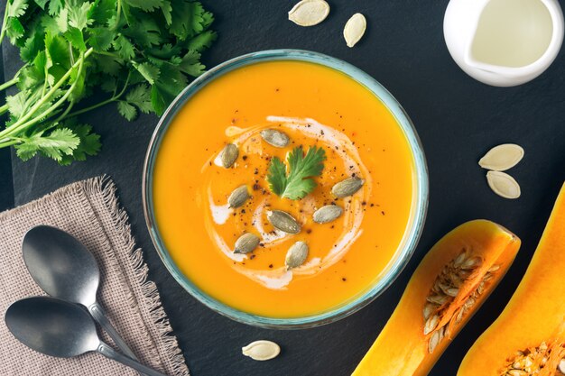 Butternut squash or pumpkin soup in a bowl. Ingredients, cream, cilantro, pumpkin slices and seeds on slate board. Healthy vegetarian food. Halloween, Thanksgiving diner. Copy space.