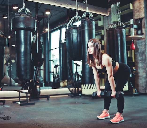 Тренировка ягодиц. Подходящая женщина в спортивной одежде делает тягу с гантелями в тренажерном зале