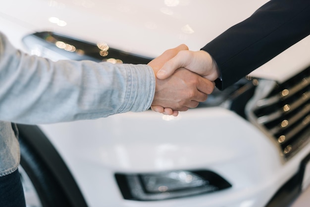 Buyer of car shaking hands with seller in auto dealership, in front of car. Close-up of handshake of unrecognizable business people. Concept of choosing and buying new car at showroom.
