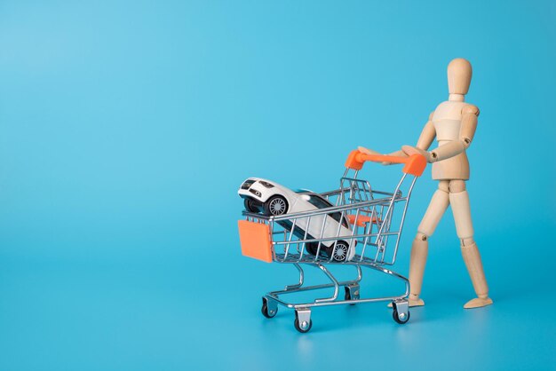 Buying new car concept. Photo of wooden male figure carrying shopping cart with mini white car inside isolated blue backdrop with copy space