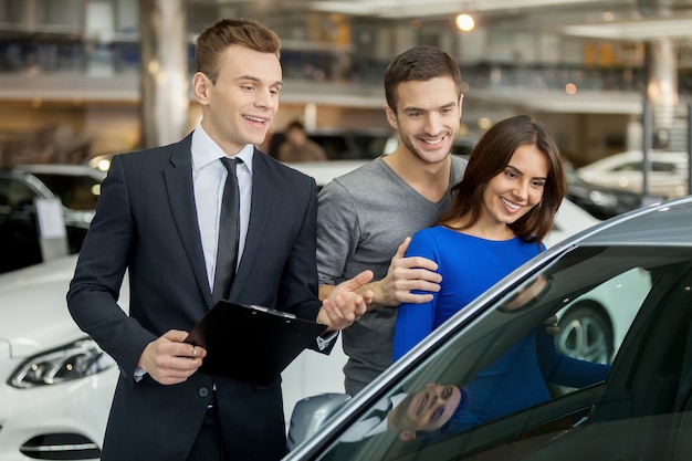 Buying their first car together. Handsome young car salesman standing at the dealership telling about the features of the car to the customers