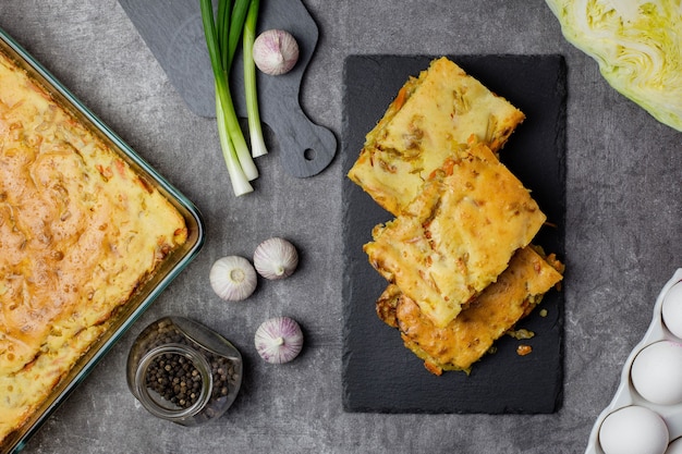 Cabbage pie with carrots on a dark concrete with ingredients for cooking, eggs, green onions, vegetables and spices.