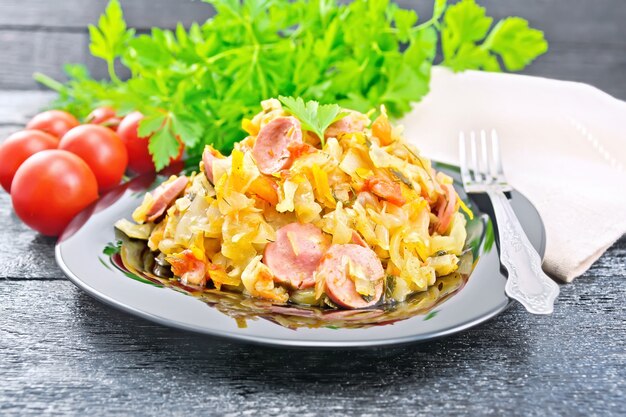 Cabbage stew with sausages in a black plate, napkin, tomatoes, parsley and fork on a wooden board