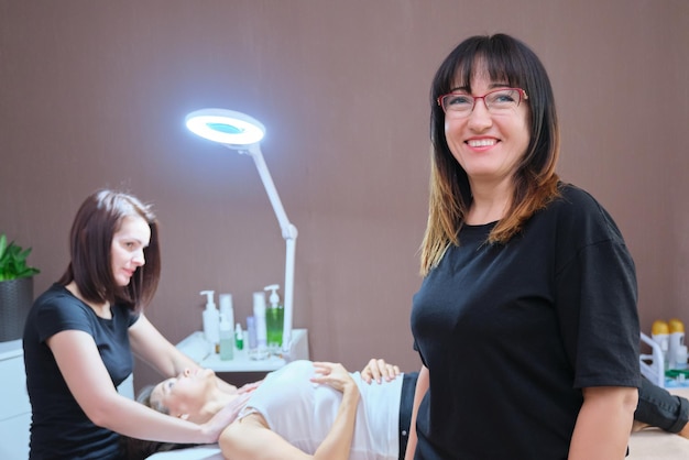 Cabinet of cosmetologist, dermatologist, massage therapist. Portrait of business confident woman owner, manager, background working masseur beautician with client