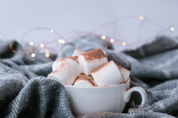 Cacaopoeder toevoegen bovenop warme chocolademelk met marshmallows in witte kop op trui achtergrond. Gezellige kerstverlichting. Voorbereiding op nieuwjaarsvakantie sfeer
