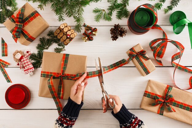 Cadeau verpakking achtergrond. Bovenaanzicht op vrouwelijke handen verpakken kerstcadeau in ambachtelijk papier, decoreren met satijnen lint op witte rommelige rustieke houten tafel. Wintervakantie concept.