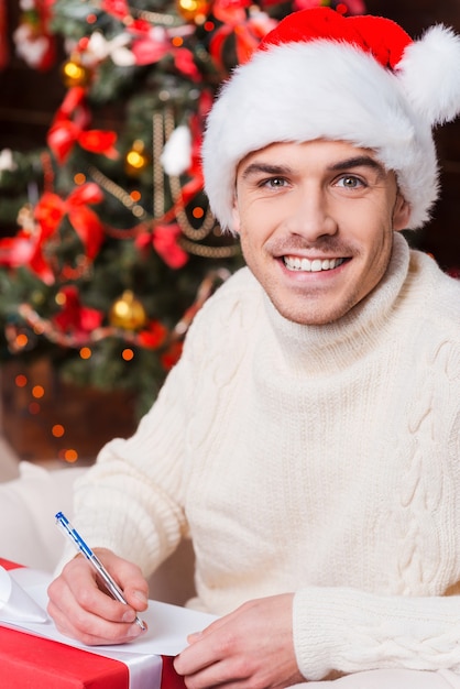 Cadeau voor haar. Knappe jonge man in kerstmuts die kerstbrief schrijft en lacht met kerstboom op de achtergrond