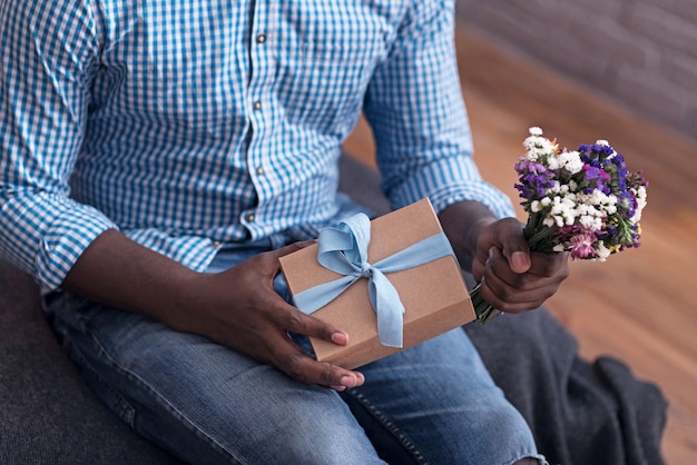 Cadeau voor jou. Close up van opgetogen man met bloemen en cadeau en verrassing voorbereiden voor zijn vriendin.