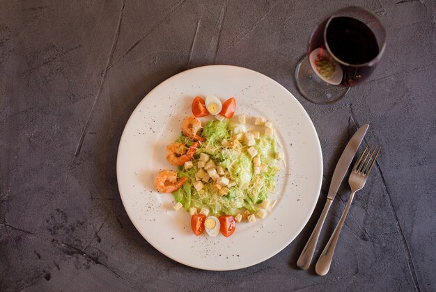 Caesar salad with shrimp on the white plate, glass of wine and fork and knife on the grey textured background. Restourant menu. Server example.